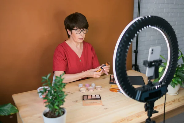 Beauty and cosmetics e-commerce and blogging concept - professional mature female make up artist testing new cosmetics recording video review on cosmetic products at home