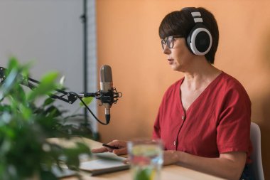 Mature woman making podcast recording for her online show. Attractive business woman using headphones front of microphone for a radio broadcast