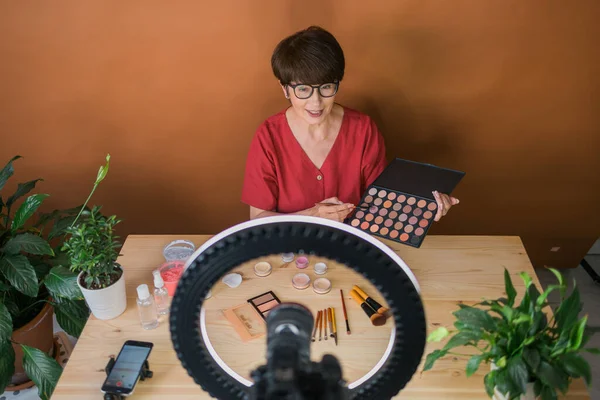 Middle-aged woman talking on cosmetics with makeup eye shadows and blush palette and brushes while recording her video. Mature female making video for her blog on cosmetics