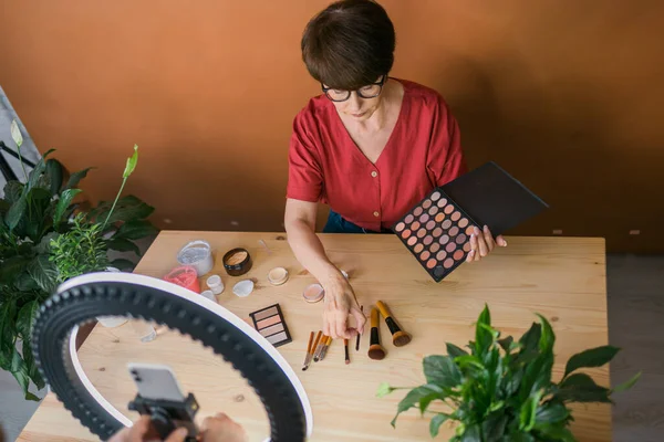 Beauty and cosmetics e-commerce and blogging concept - professional mature female make up artist testing new cosmetics recording video review on cosmetic products at home