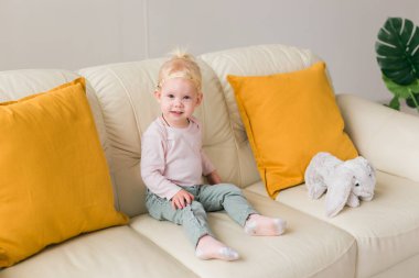 Little girl with cochlear implant sitting on sofa at home. Hear impairment and deaf community concept