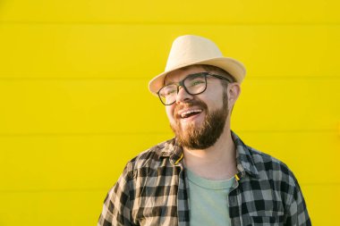 Portrait positive young hipster man smiling over yellow wall store, outdoor background - Handsome trendy bearded guy with hat and glasses standing outdoors - Fashion and trendy youth people