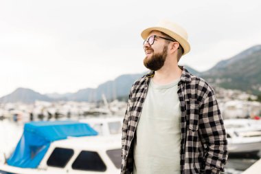 Handsome man wearing hat and glasses near marina with yachts. Portrait laughing man with sea port background with copy space