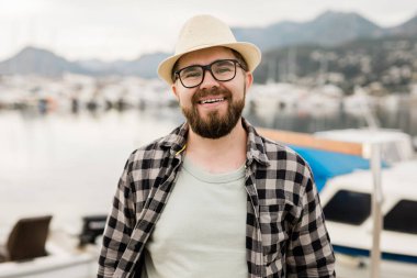 Handsome man wearing hat and glasses near marina with yachts. Portrait laughing man with sea port background with copy space