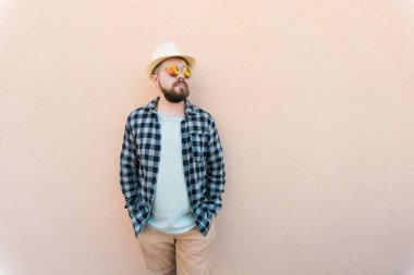 Yazlık şapka ve ekose gömlek giyen yakışıklı beyaz adam duvarın yanında mutlu bir şekilde gülümsüyor.