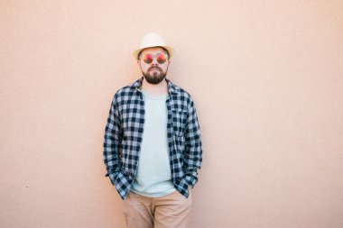 Yazlık şapka ve ekose gömlek giyen yakışıklı beyaz adam duvarın yanında mutlu bir şekilde gülümsüyor.