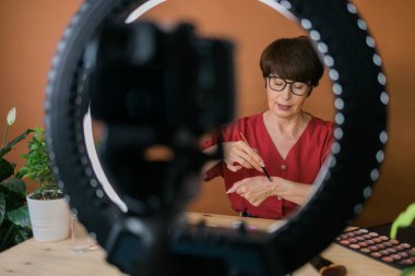 Middle-aged woman talking on cosmetics with makeup eye shadows and blush palette and brushes while recording her video. Mature female making a video for her blog on cosmetics