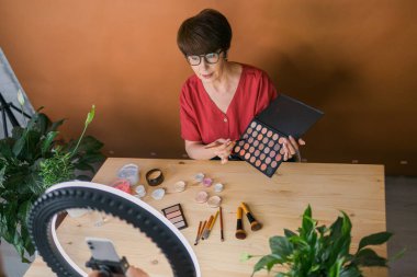 Middle-aged woman talking on cosmetics with makeup eye shadows and blush palette and brushes while recording her video. Mature female making a video for her blog on cosmetics