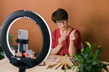 Middle-aged woman talking on cosmetics with makeup eye shadows and blush palette and brushes while recording her video. Mature female making a video for her blog on cosmetics
