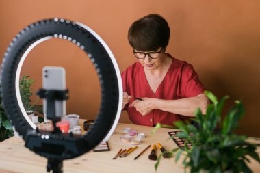 Middle-aged woman talking on cosmetics with makeup eye shadows and blush palette and brushes while recording her video. Mature female making a video for her blog on cosmetics