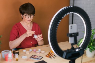 Middle-aged woman talking on cosmetics with makeup eye shadows and blush palette and brushes while recording her video. Mature female making a video for her blog on cosmetics