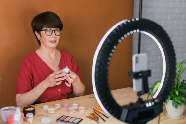 Beauty and cosmetics e-commerce and blogging concept - professional mature female make up artist testing new cosmetics recording video review on cosmetic products home