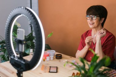 Middle-aged woman talking on cosmetics with makeup eye shadows and blush palette and brushes while recording her video. Mature female making a video for her blog on cosmetics