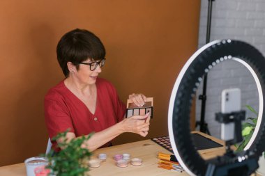 Beauty and cosmetics e-commerce and blogging concept - professional mature female make up artist testing new cosmetics recording video review on cosmetic products home