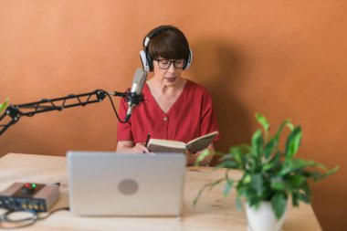 Mature woman making podcast recording for her online show. Attractive business woman using headphones front of microphone for a radio broadcast.
