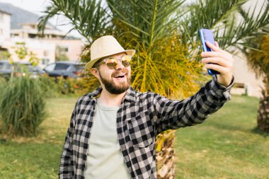 Man taking selfie portrait over palm tree background - Happy millennial guy enjoying summer holidays in city - Youth and technologies.