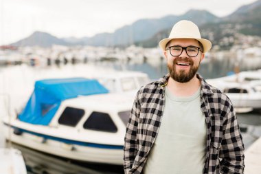 Handsome man wearing hat and glasses near marina with yachts. Portrait laughing man with sea port background with copy space