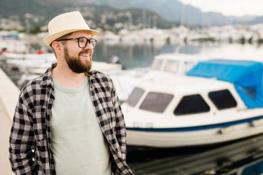 Handsome man wearing hat and glasses near marina with yachts. Portrait laughing man with sea port background with copy space