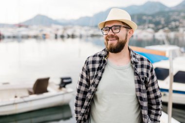 Handsome man wearing hat and glasses near marina with yachts. Portrait laughing man with sea port background with copy space