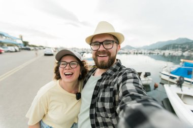Young couple taking a self portrait laughing as they pose close together for camera on their smartphone outdoors in summer port with boats and yachts.