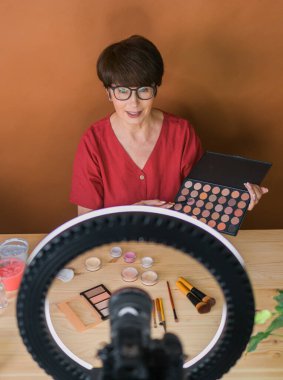 Middle-aged woman talking on cosmetics with makeup eye shadows and blush palette and brushes while recording her video. Mature female making a video for her blog on cosmetics