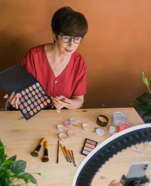 Middle-aged woman talking on cosmetics with makeup eye shadows and blush palette and brushes while recording her video. Mature female making a video for her blog on cosmetics