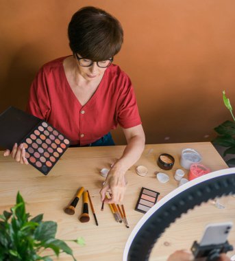 Beauty and cosmetics e-commerce and blogging concept - professional mature female make up artist testing new cosmetics recording video review on cosmetic products home