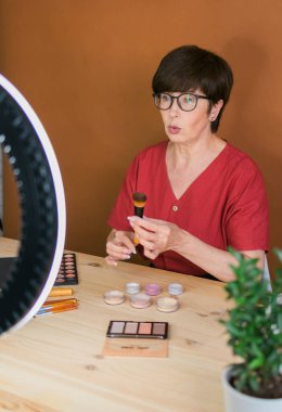 Beauty and cosmetics e-commerce and blogging concept - professional mature female make up artist testing new cosmetics recording video review on cosmetic products home