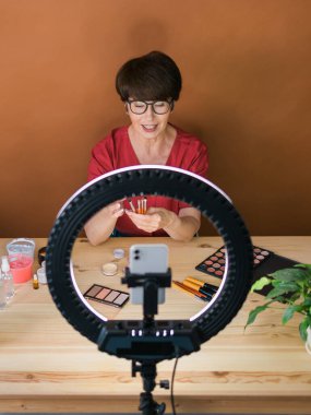 Middle-aged female beauty blogger or influencer with makeup product recording video in room at home. Using ring lamp