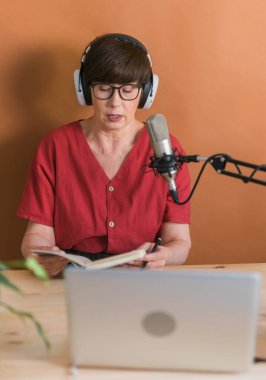 Mature woman making podcast recording for her online show. Attractive business woman using headphones front of microphone for a radio broadcast.