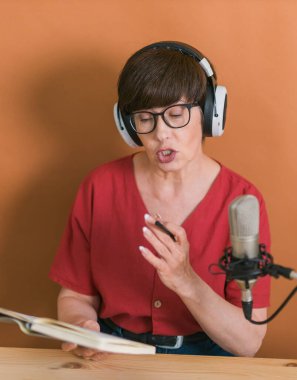 Mature woman making podcast recording for her online show. Attractive business woman using headphones front of microphone for a radio broadcast.