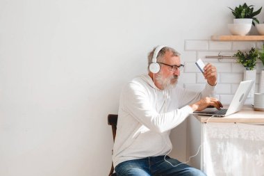 Üst düzey yaşlı adam dizüstü bilgisayarla online ödeme yapıyor. Gülümseyen yaşlı büyükbaba kredi kartıyla alışveriş yapıyor. Emeklinin elinde internet bankacılığı için banka kartı var