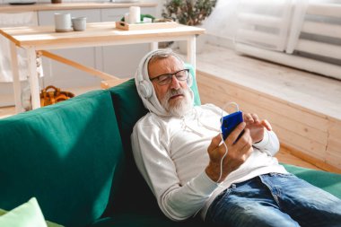 Son sınıf hipster 'ı cep telefonuyla mobil uygulama üzerinden müzik dinlerken kullanıyor. Yaşlı erkek video akıllı telefon izlerken yüz koruyucu maske takıyor.