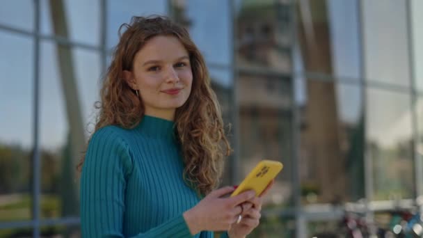 Portrait de jeune femme aux cheveux blonds bouclés surfant sur Internet sur smartphone et regardant la caméra debout sur la rue de la ville. Concept de personnes, gadgets modernes et modes de vie. — Video