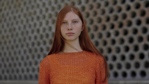 街の通りに一人で立っている赤い髪とそばかすを持つかなり白人女性。カメラを見てオレンジ色のニットセーターの若い女性の屋外の肖像画。ライフスタイルと若者のコンセプト. — ストック動画
