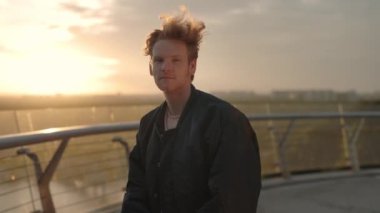 Kırmızı kıvırcık saçlı, gülümseyen genç adamın portresi kameraya bakıyor. Akıllı telefonu elinde tutuyor ve gün batımında dışarıda boş vakit geçiriyor..