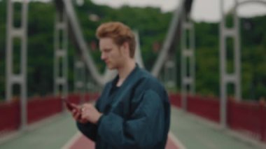 Şık giyinmiş genç bir adamın portresi elinde akıllı bir telefonla şehir köprüsünde duran kameraya bakıyor. Dışarıda zaman geçirmek. Modern aletleri kullanarak..