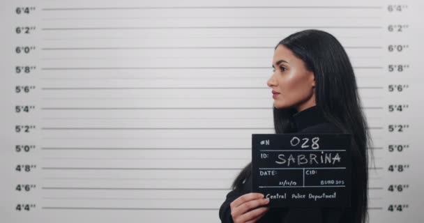 Side profile mugshot of female mixed race person with long hair turning head and looking to camera. Criminal attractive woman with sign for photo in front of police metric lineup wall. — Stock Video
