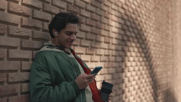 Стильный парень в наушниках пьет кофе и пользуется мобильным телефоном — стоковое видео