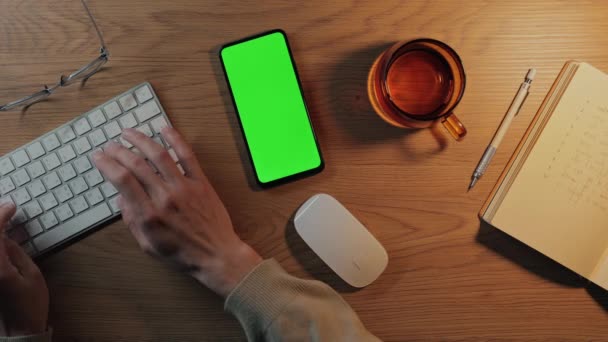 Человек сидит за столом с клавиатурой и зеленым экраном мобильного — стоковое видео