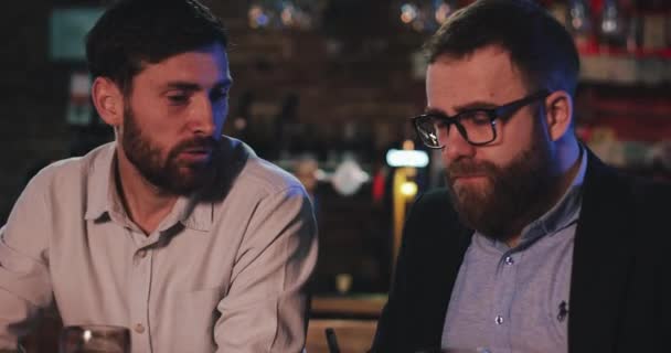 Primer plano de alegres amigos varones conversando mientras beben cerveza en el pub. Dos hombres relajándose después del día de trabajo, hablando y sonriendo mientras están sentados en el bar. Concepto de ocio. — Vídeos de Stock