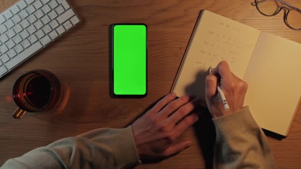 Desplazamiento del hombre en el móvil de pantalla verde y escritura en planificador — Vídeo de stock