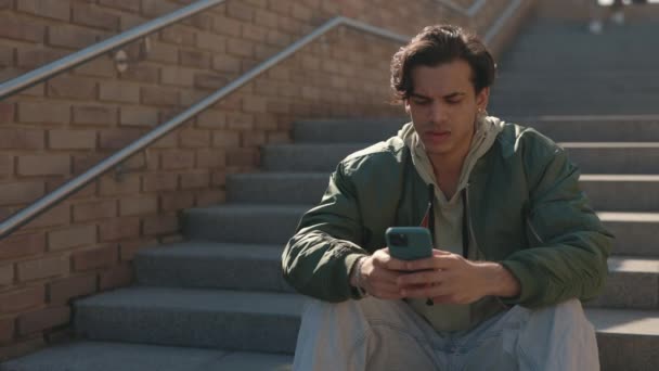 Стильный парень пишет смс на смартфоне, сидя на улице — стоковое видео