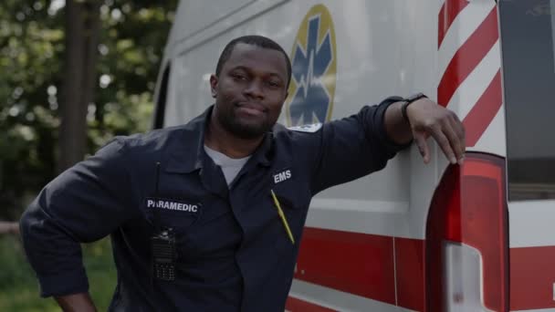 Позитивный африканский американский парамедик позирует возле скорой помощи на открытом воздухе — стоковое видео