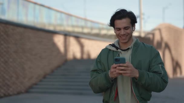 Улыбающийся мужчина в наушниках ходит с мобильным в руках — стоковое видео