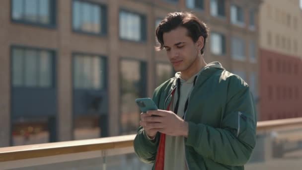 Hombre sonriendo en la cámara mientras está de pie en la calle con el móvil — Vídeos de Stock