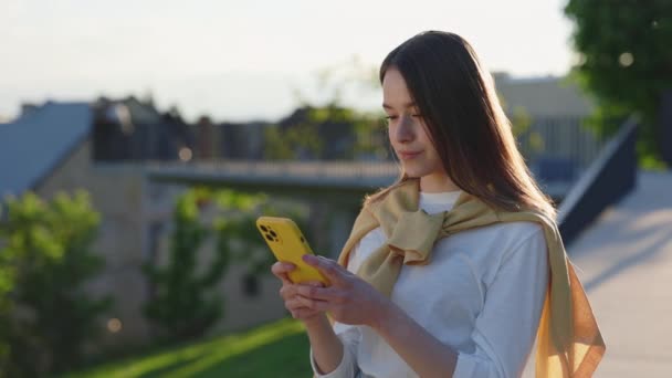 Retrato de mulher jovem com cabelo castanho em pé na rua e mensagens de digitação no smartphone. Mulher caucasiana usando gadget moderno para conversar ao ar livre e olhar para a câmera. — Vídeo de Stock