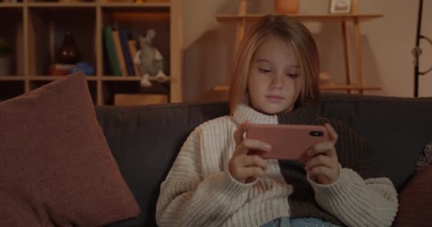 Crop view of pretty girl watching video while sitting on sofa at home.Young child with long blond hair holding smartphone horizontally while looking and touching screen. — стоковое видео