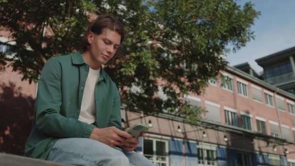 Handsome man texting on smartphone while sitting outdoors — Stock Video