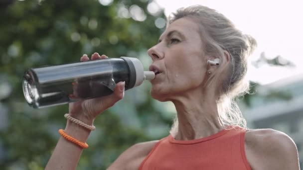 Mujer envejecida en ropa deportiva bebiendo agua después de correr — Vídeos de Stock